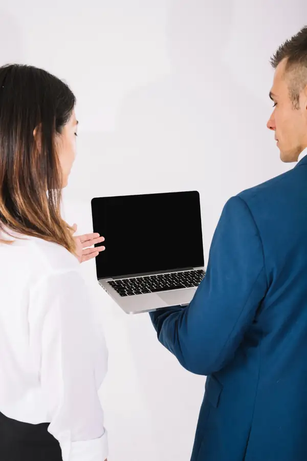 A consultant in a blue suit holding out an open laptop to a colleague, seen from behind.