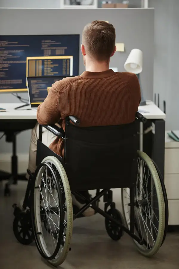 Seen from behind, a person in a wheelchair working at a desk with code on two screens.