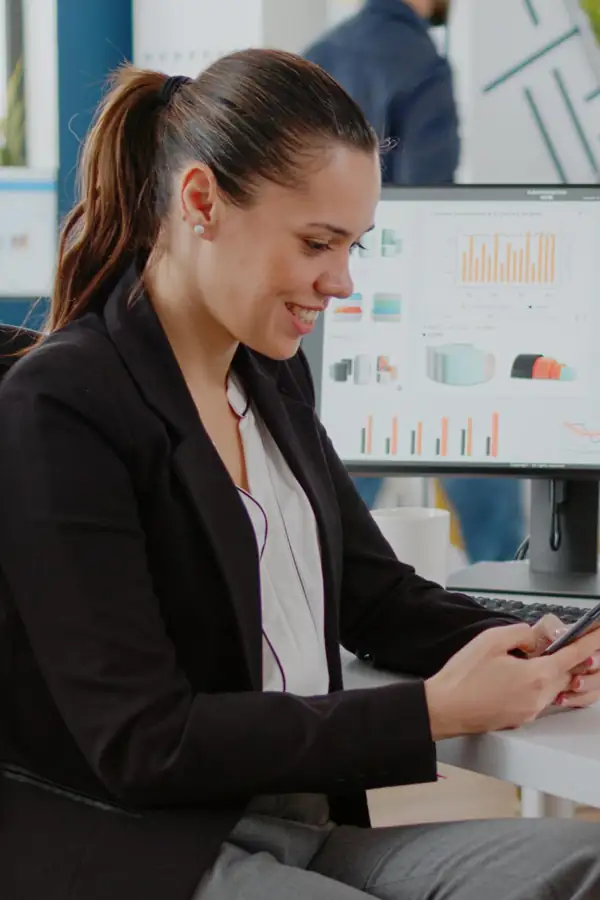 A shop manager checking her phone at a desk, with a store dashboard on the monitor behind her.