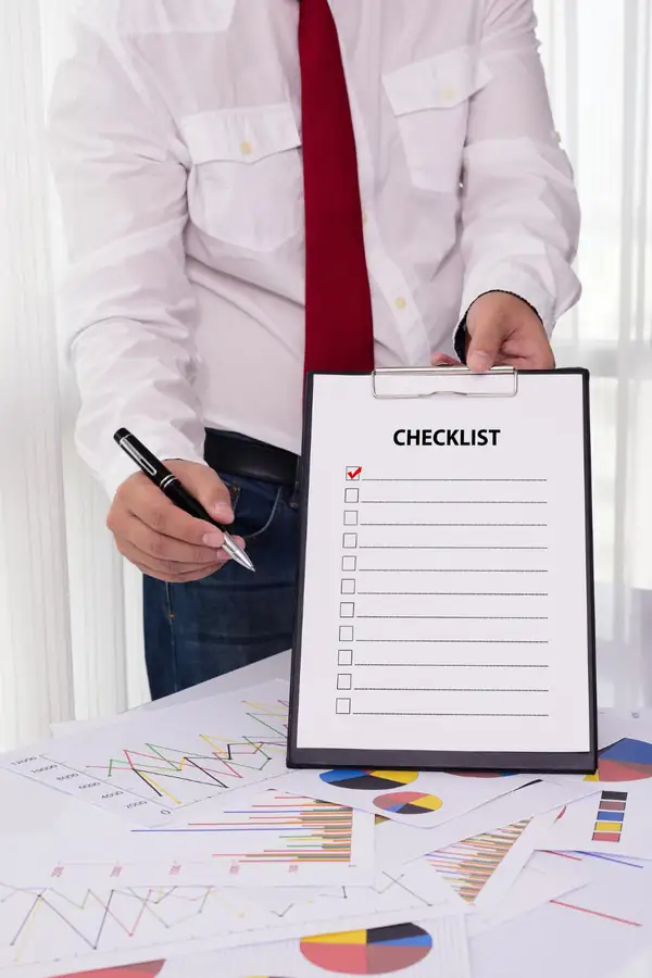 A man holding out a clipboard headed CHECKLIST with a pen, printed charts spread on the desk below.