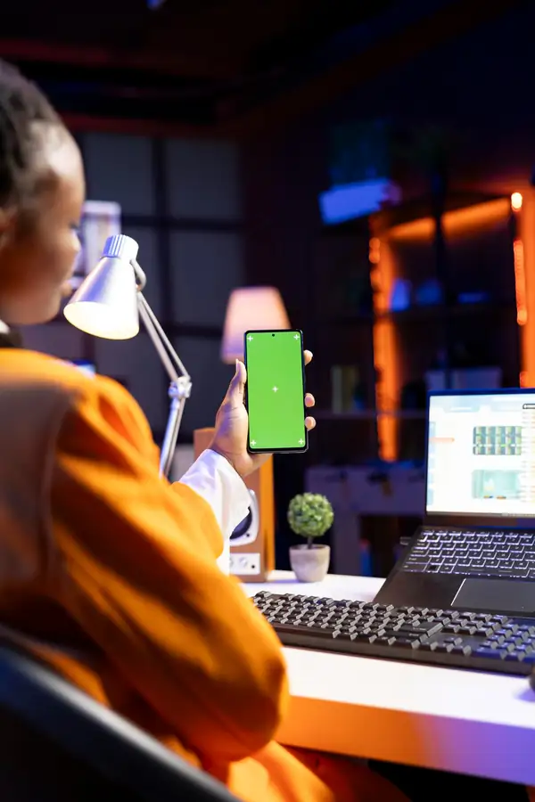 A person holding a phone beside a laptop showing a dashboard, working in the evening.