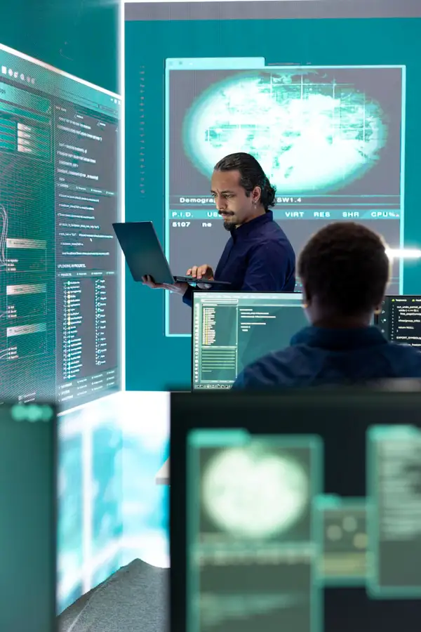 Two engineers working among large screens of code and live dashboards.