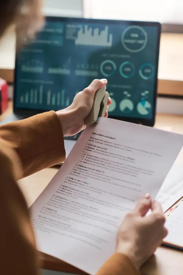 Hands stapling a printed agreement in front of a monitor showing charts.