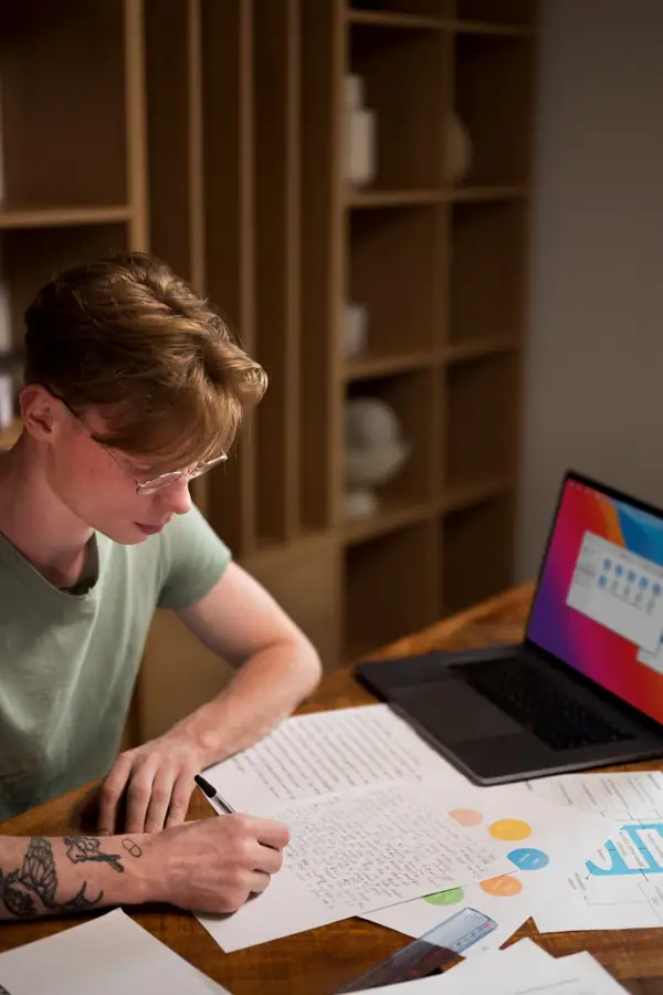 A young man writing notes by hand beside an open laptop and printed pages.