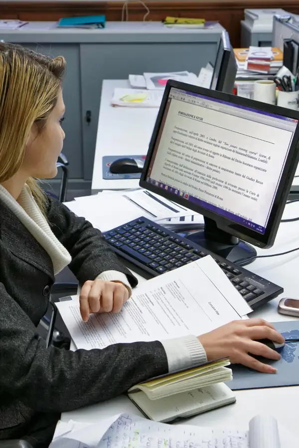 A woman at an office desk comparing a printed document with a legal file on screen.