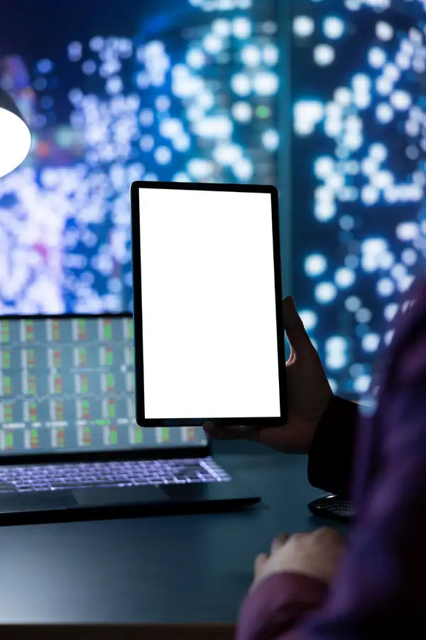 A hand holding a tablet with a completely blank white screen, data screens glowing behind.