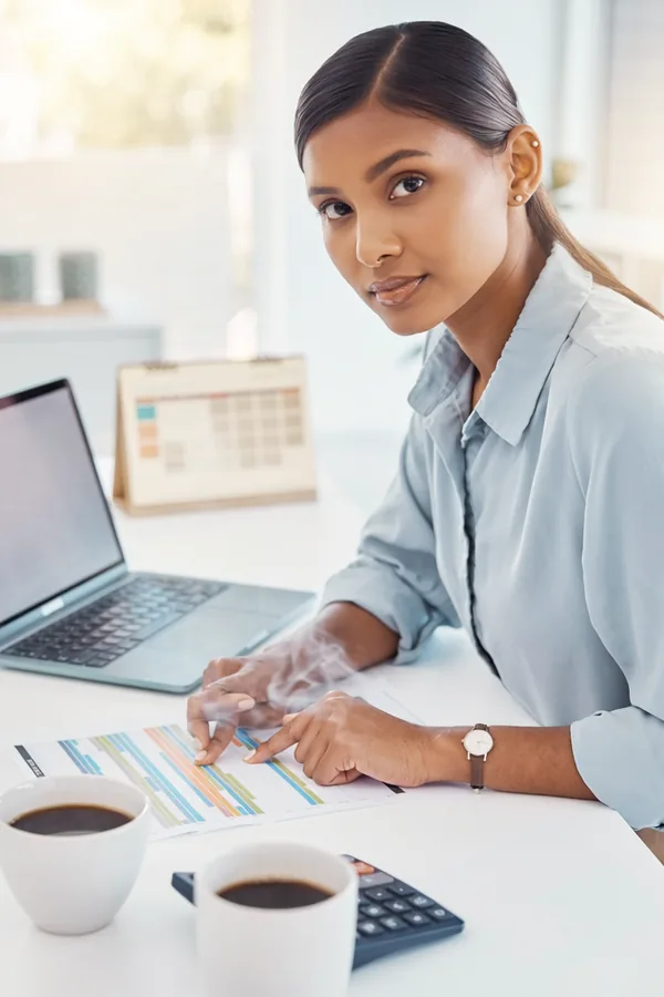A woman at a bright desk points at a printed project timeline beside a laptop, with a desk calendar and coffee cups nearby.