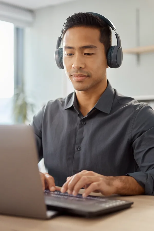 A man wearing over-ear headphones types on a laptop at a desk by a window, listening as he works.