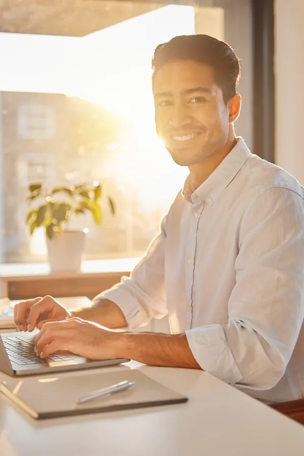 A smiling man in a light shirt types on a laptop at a tidy desk, with a plant and warm afternoon light behind him.