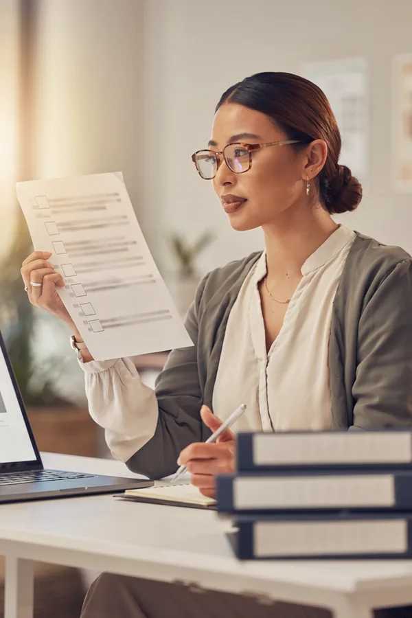 A woman in glasses holds up a printed checklist and compares it with a website open on her laptop, binders stacked beside her.