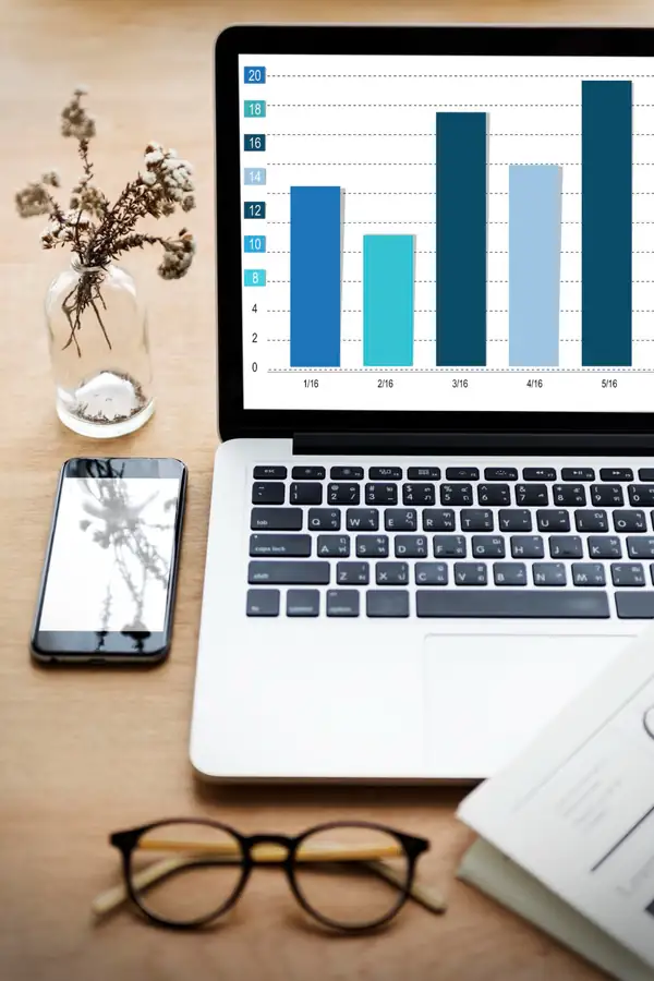 A laptop on a desk showing a bar chart, with a phone, glasses and a printed report beside it.