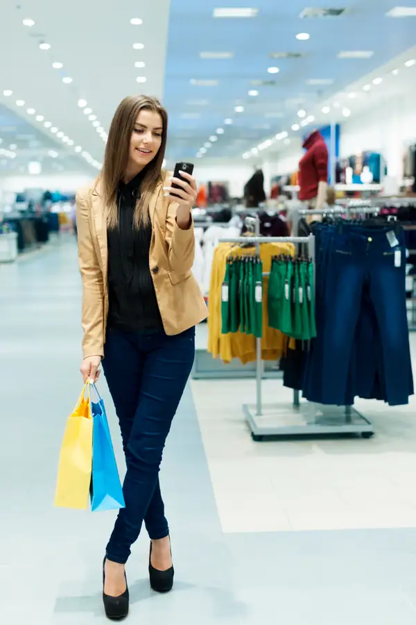 A shopper walking through a clothing store looking at her phone, carrying bags.