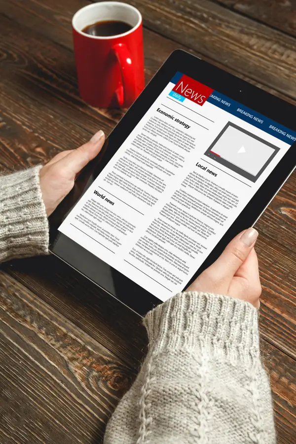 Hands holding a tablet showing a news page laid out in columns with headings and an article.