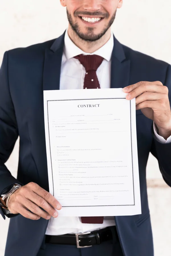 A man in a navy suit and red tie holds up a printed contract with both hands.