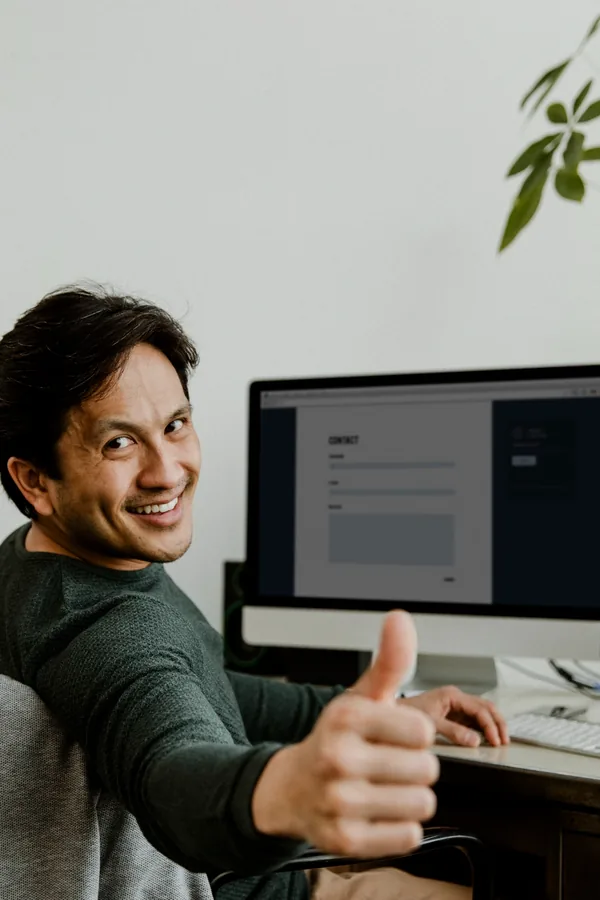 A man at a desk turns to give a thumbs up, with a website contact form open on the monitor behind him.