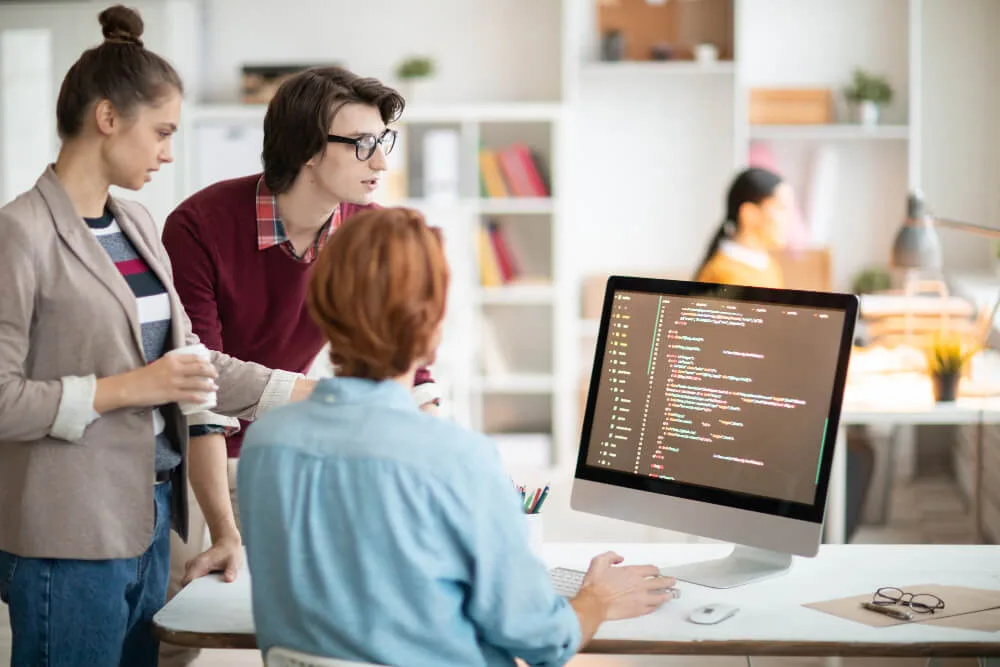 Three developers gathered at a desk look at lines of code on a large monitor while discussing a fix.