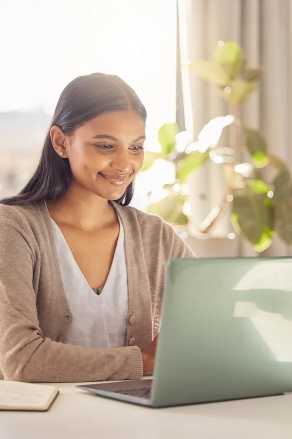 A woman in a cardigan smiles as she reads something on her laptop at a desk by a bright window, a plant beside her.