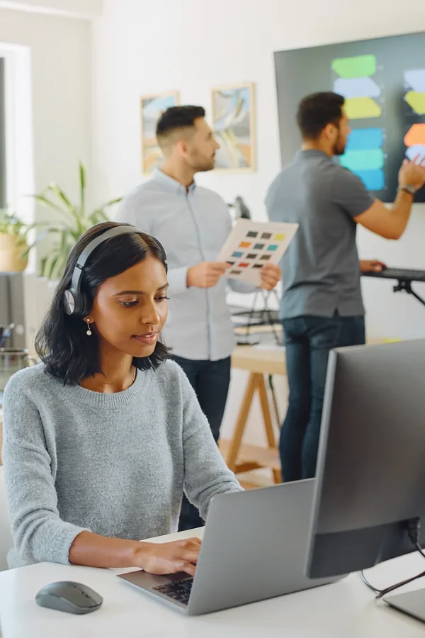 In a bright studio a woman wearing headphones tests a website on a laptop while two colleagues discuss a design pinned to a board behind her.