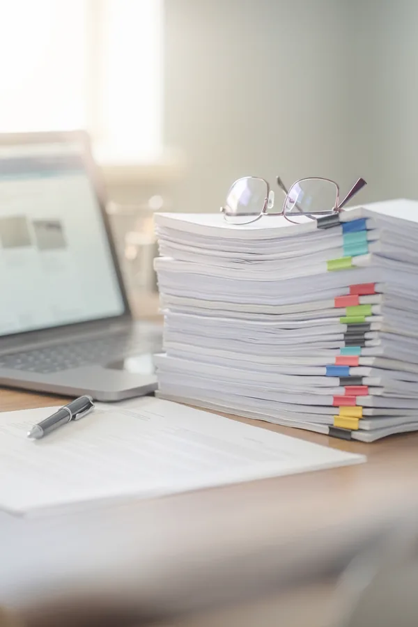 A tall stack of printed documents with coloured index tabs sits on a desk beside an open laptop, a pair of reading glasses resting on top.