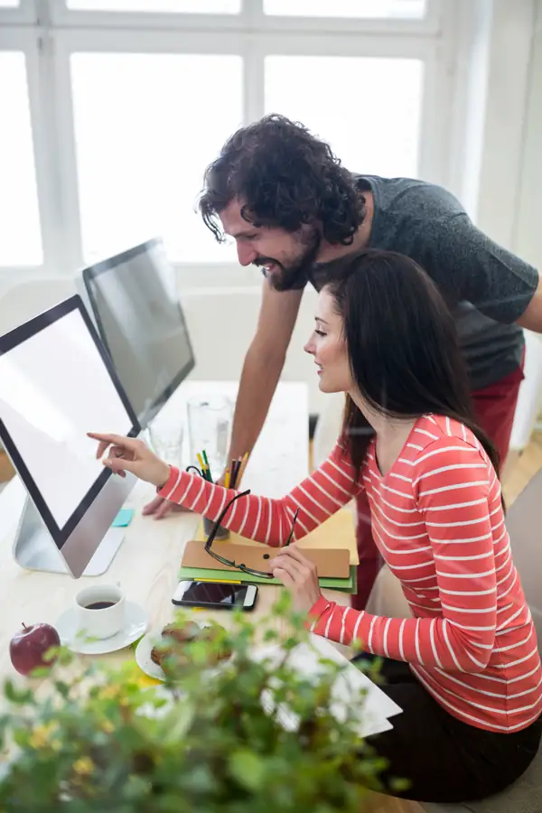 Two colleagues at a monitor, one pointing at a clean empty screen, both smiling.