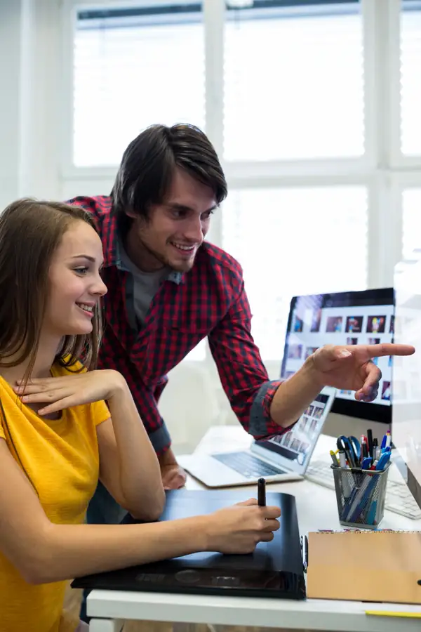 Two designers at a desk looking at a laptop showing a grid of images, one pointing at the screen.