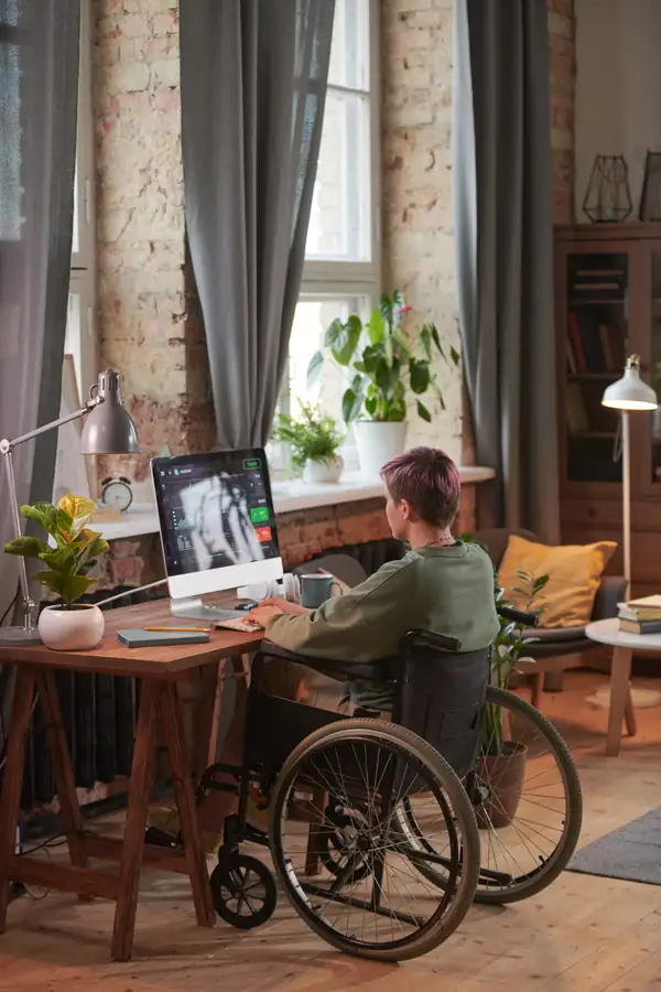 A person in a wheelchair working at a desktop computer in a bright home office.