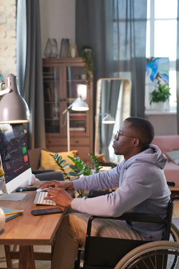 A man in a wheelchair working at a desktop computer at home, focused on the screen.